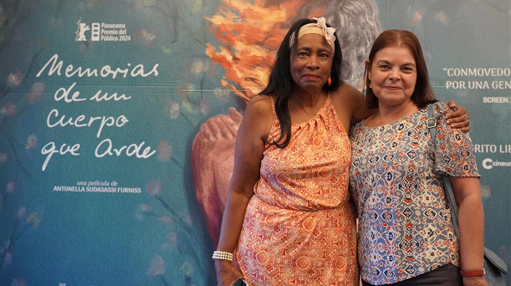 Dos mujeres abrazadas sonriendo con el banner de Memorias de un cuerpo que arde de fondo