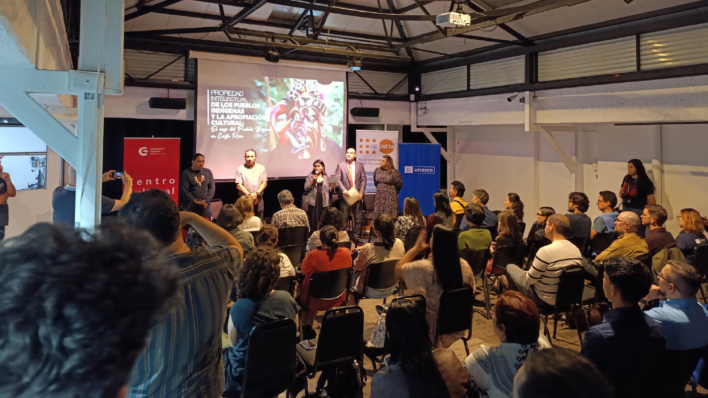 Personas participantes del conversatorio, con el panel de pie al frente del escenario.
